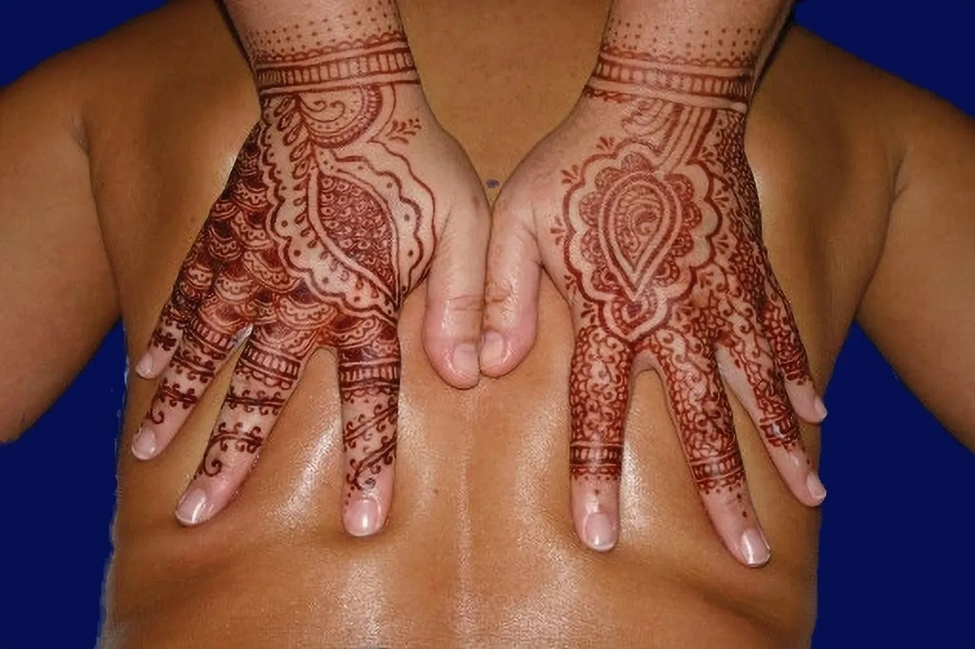 The backs of two hands covered in detailed reddish-brown henna patterns, resting on a person's back